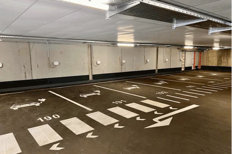 Leeres, beleuchtetes Parkdeck mit nummerierten Stellflaechen, Pfeilen und Zebrastreifen in einer Tiefgarage.