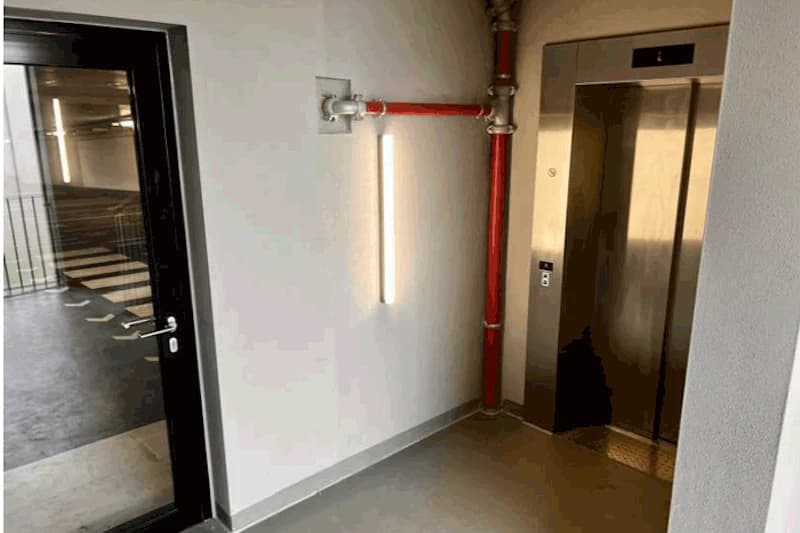 Fahrstuhltür, rote Rohrleitung, Leuchte an weißer Wand und Glastür zum Parkdeck mit Fahrbahnmarkierung.