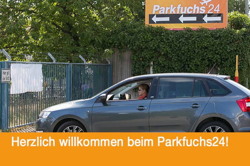 Silbernes Auto mit Fahrerin neben eingezäuntem Tor und Parkfuchs24-Schild im Hintergrund.