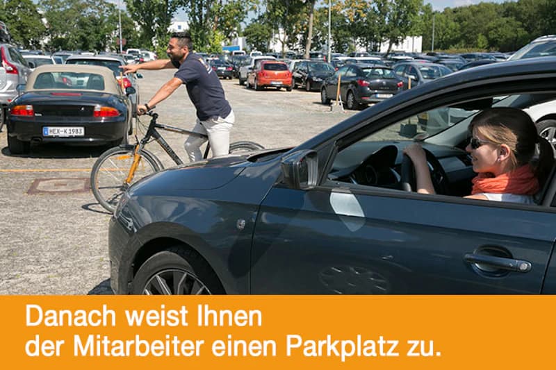 Frau sitzt im Auto und schaut, ein Mann mit Fahrrad zeigt auf einem vollen Parkplatz Richtung.