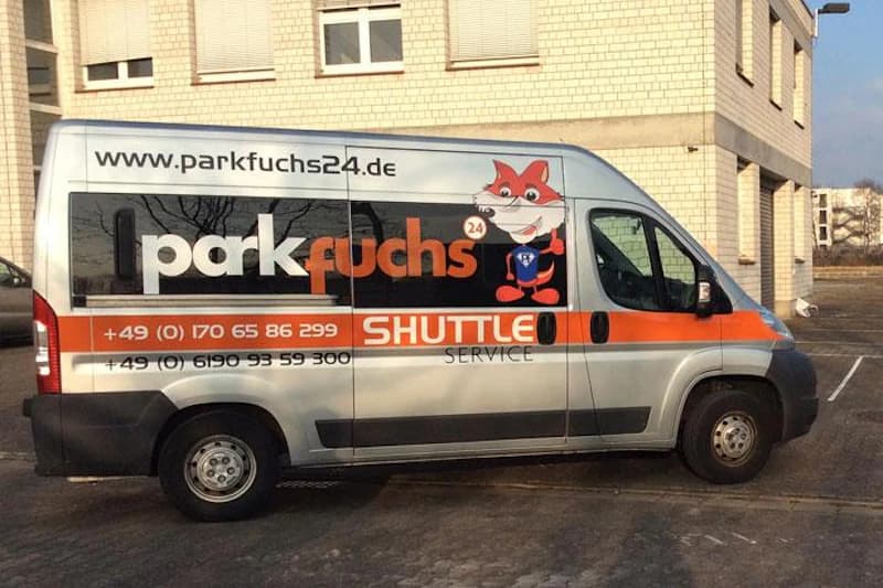 Weißer Transporter mit Parkfuchs24-Logo, Fuchs-Maskottchen und Telefonnummern vor beigem Gebäude.