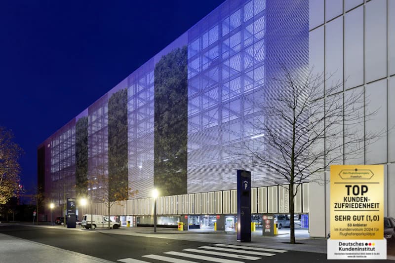 Mehrstöckiges Parkhaus mit Metallfassade und beleuchteter Einfahrt, Zebrastreifen und Lieferfahrzeugen bei Nacht.
