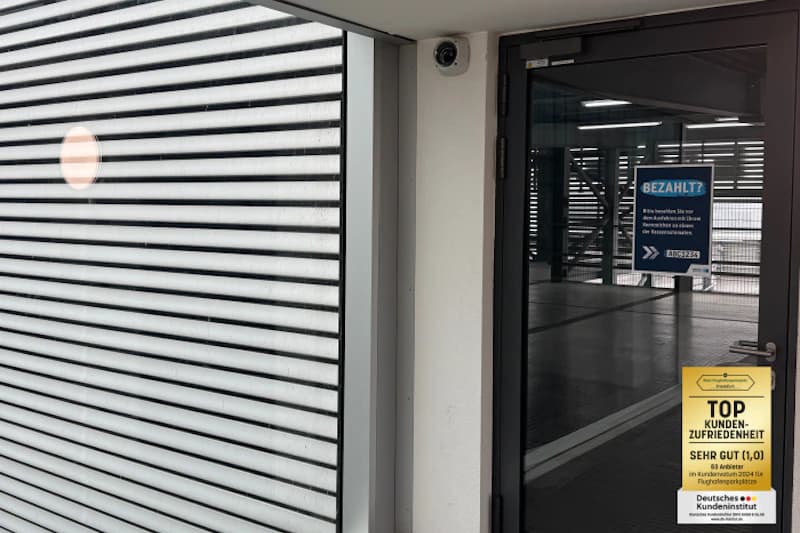 Glastür zu Parkhaus mit Sicherheitskamera über der Tür und geschlossenem Metallrollladen links.