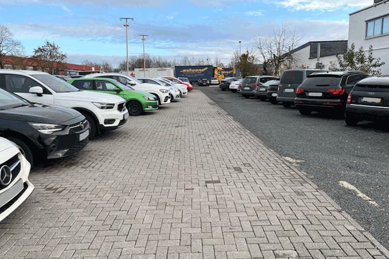 Parkplatz mit geparkten Autos in zwei Reihen, gepflasterter Mittelweg und Lkw im Hintergrund.