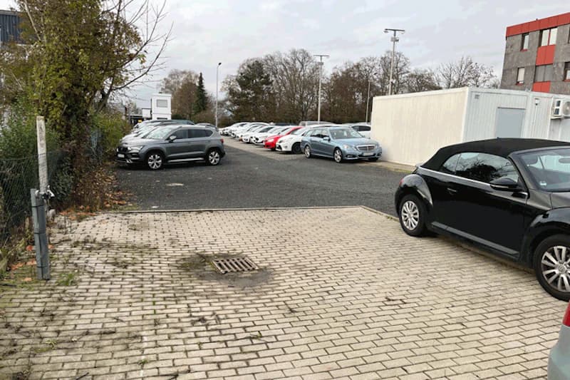 Parkplatz mit Reihen geparkter Autos, gepflasterte Fläche im Vordergrund, Container und Zaun.