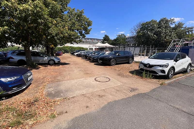 Offener Schotterparkplatz mit mehreren geparkten Autos, Bäumen, Zaun und Gebäuden im Hintergrund.