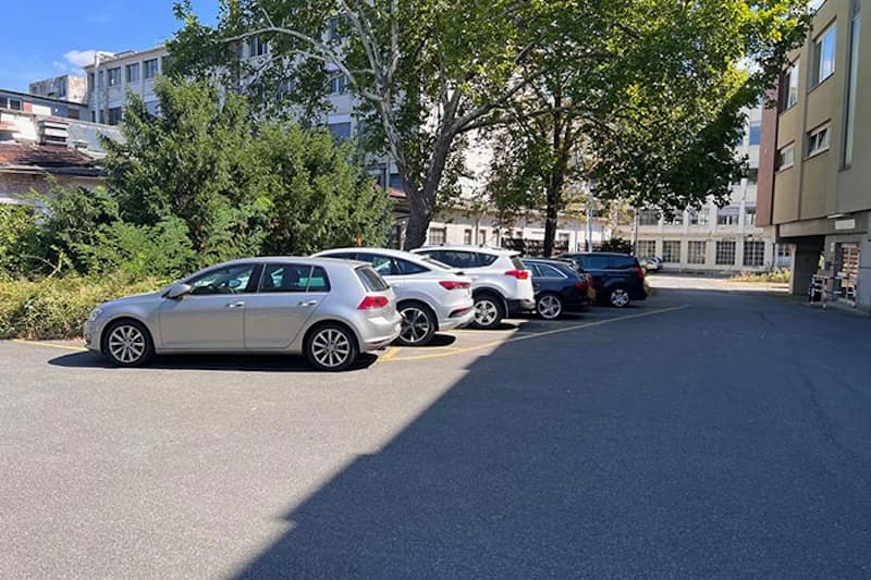 Sechs geparkte Autos in einer Außenreihe neben Gebäuden und Bäumen, Sonnenschein und Schatten auf dem Asphalt.
