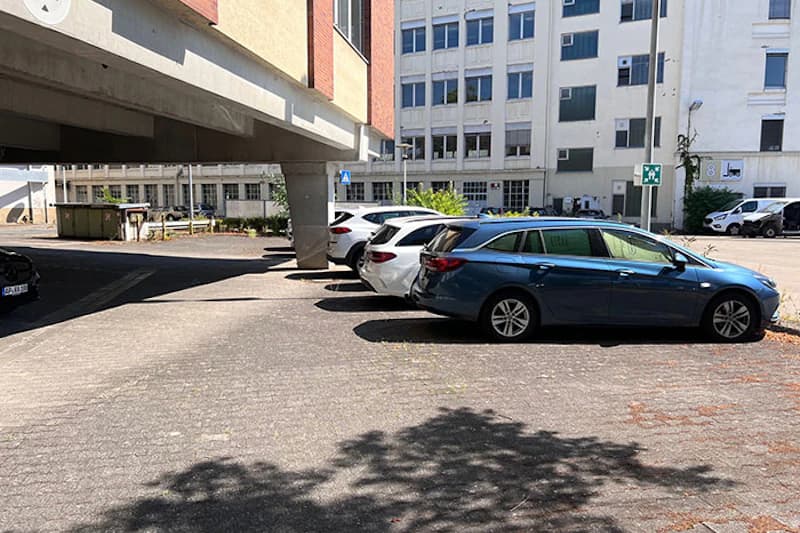 Außenparkplatz neben Bürogebäude mit drei geparkten Autos, Betonstützen, Abfallcontainer im Hintergrund und Schatten von Bäumen.