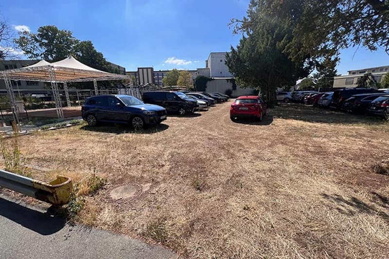 Trockenes Grasfeld mit geparkten Autos, Pavillon links und Gebäuden im Hintergrund.
