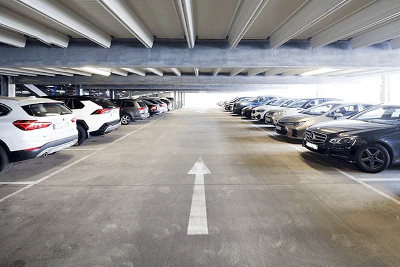 Beleuchtetes Parkhaus mit mittiger Fahrspur und weißem Richtungspfeil, geparkte Autos links und rechts.