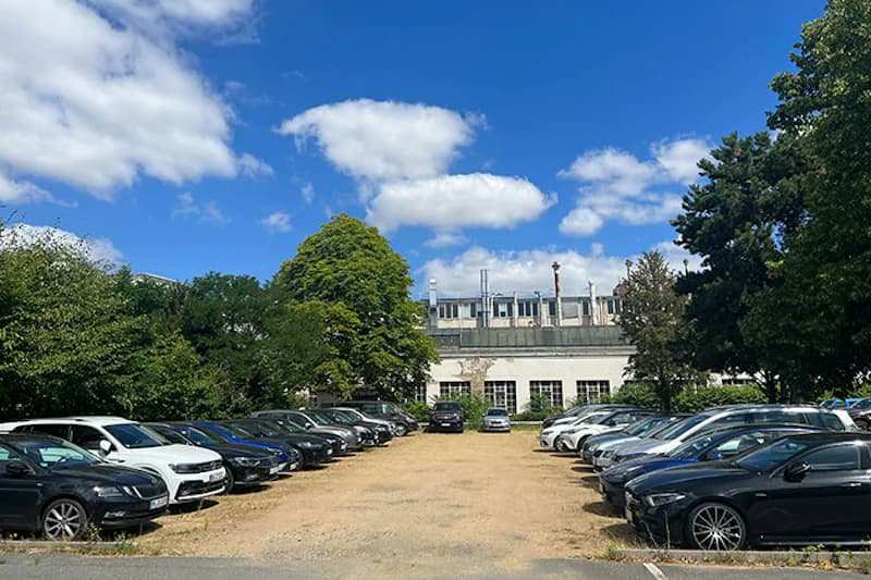 Befestigter Parkplatz mit zwei Reihen geparkter Autos, Gebäude im Hintergrund und Bäume unter blauem Himmel.