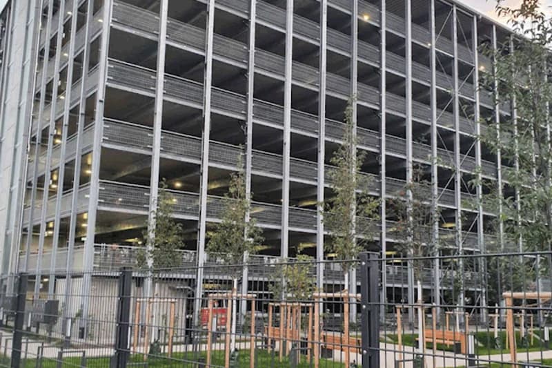 Mehrgeschossiges offenes Parkhaus mit Stahlkonstruktion, beleuchteten Ebenen und Zaun im Vordergrund.