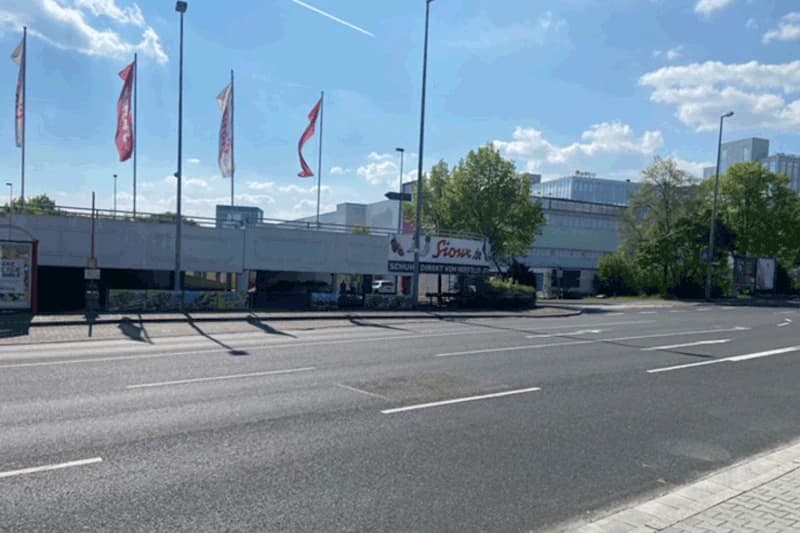 Mehrstöckiges Parkdeck neben leerer Straße, Fahnen auf Masten und Buswartehäuschen bei sonnigem Himmel.