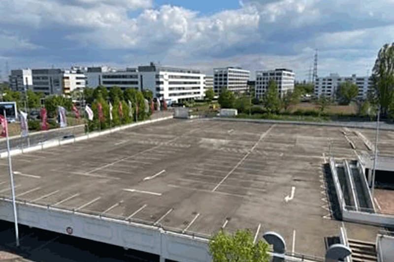 Leeres oberes Parkdeck mit markierten Stellplätzen, Treppe rechts und Bürogebäuden im Hintergrund.