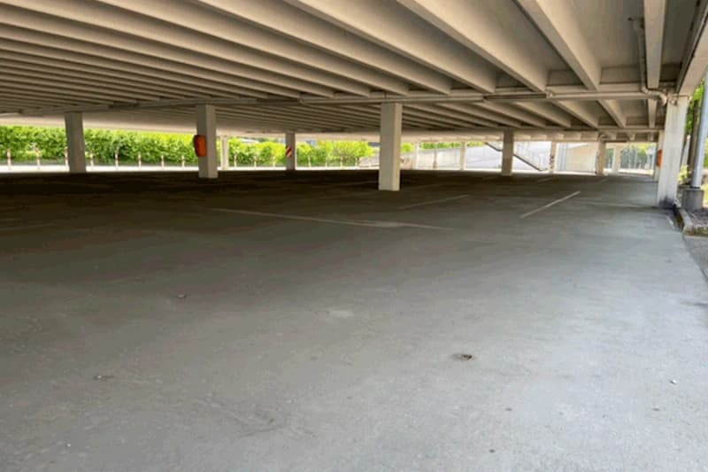 Leeres überdachtes Parkdeck mit Stützpfeilern, sichtbaren Deckenbalken und markierten Parkbuchten, Hecke und Zaun im Hintergrund.