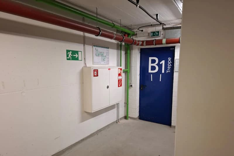 Gang in Tiefgarage mit blauer Tür beschriftet B1 Treppe, weißem Feuerlöschschrank, grünem Notausgangssymbol und roten Rohrleitungen.