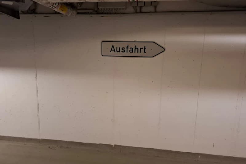Weiße Betonwand in Tiefgarage mit Pfeilschild Ausfahrt rechts und Rohren an der Decke.