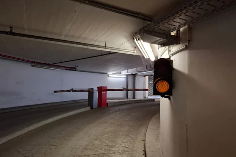 Schlauchförmige Tiefgaragenrampe mit geöffneter Schranke und gelbem Verkehrslicht an der Wand.