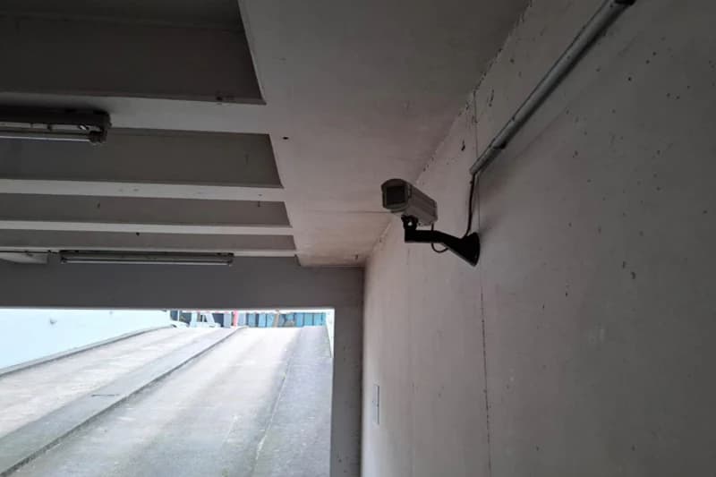 Überdachte Ausfahrt einer Tiefgarage mit an der Wand montierter Überwachungskamera und Rampe ins Freie.