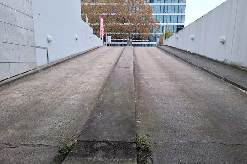 Schräge Fahrzeugzufahrt mit mittigem Betonstreifen, seitlichen Betonwänden und Gebäuden im Hintergrund.