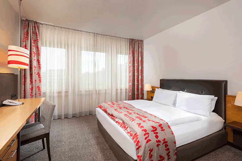 Helles Hotelzimmer mit Doppelbett, gepolstertem Kopfteil, Nachttischen, Schreibtisch und transparenten Gardinen.