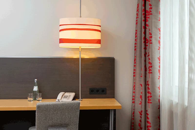 Schreibtisch mit leuchtender Tischlampe, Telefon, Wasserflasche, zwei Gläsern und gepolstertem Stuhl.