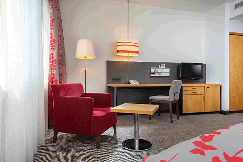 Roter Sessel neben kleinem Tisch, Steh- und Hängelampe, Schreibtisch mit Stuhl und Fernseher, Vorhang und Bettkante.