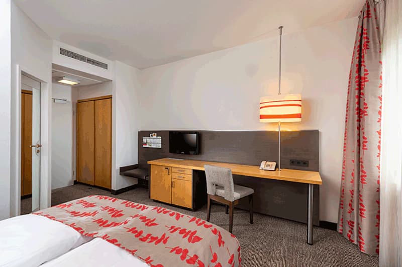 Hotelzimmer mit Doppelbett, Schreibtisch, Stuhl, Wandfernseher, Lampe, Kleiderschrank und Gardinen.