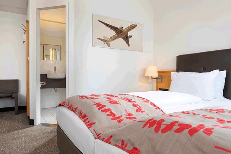 Hotelzimmer mit Bett und rot gemusterter Decke, Nachttischlampe, offenem Bad mit Waschbecken und Flugzeugbild.