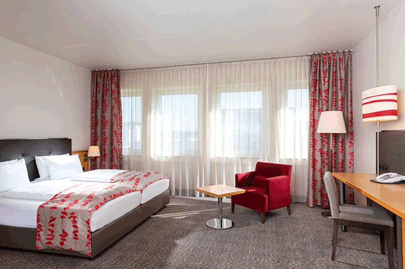 Hotelzimmer mit zwei Betten, roten gemusterten Vorhängen und Überwürfen, rotem Sessel, Schreibtisch und großen Fenstern.