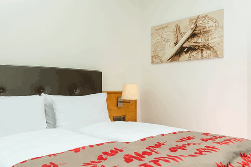 Doppelbett mit weißen Kissen, dunklem Kopfteil, Nachttischlampe und Wandbild mit Flugzeug.