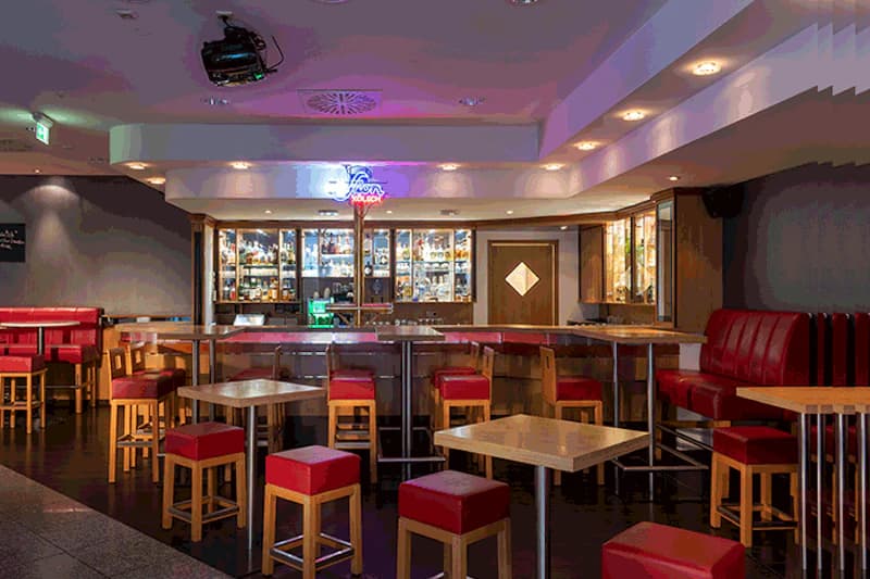 Hotelbar mit Holztheke, Rückwandregalen mit Flaschen, roten Hockern und Sitzbänken.