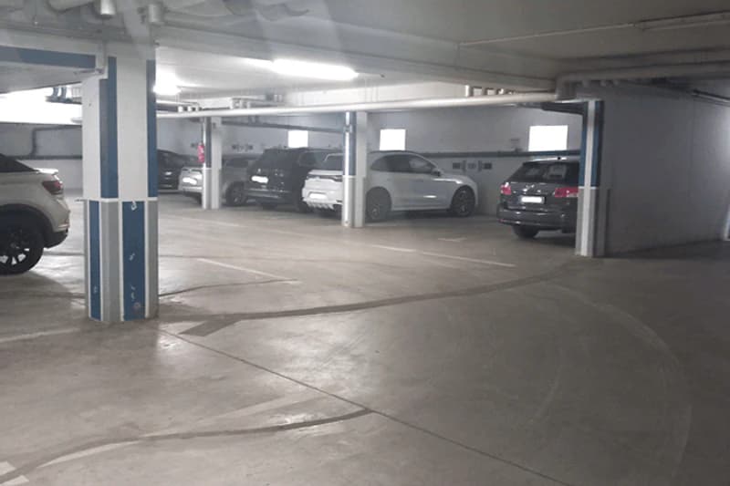 Beleuchtetes Parkdeck in Tiefgarage mit abgestellten Autos und Betonpfeilern mit blauen Markierungen.