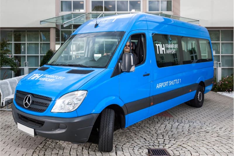 Blauer NH Frankfurt Airport Shuttlebus mit Mercedes-Logo, Fahrer am Steuer, geparkt vor Glasfassade.
