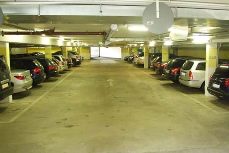 Beleuchtete Tiefgarage mit Fahrspur und geparkten Autos links und rechts, Stützen mit Nummern sichtbar.