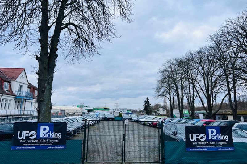Eingezäunter Parkplatz mit Reihen geparkter Autos und UFO Parking Bannern am Eingang.