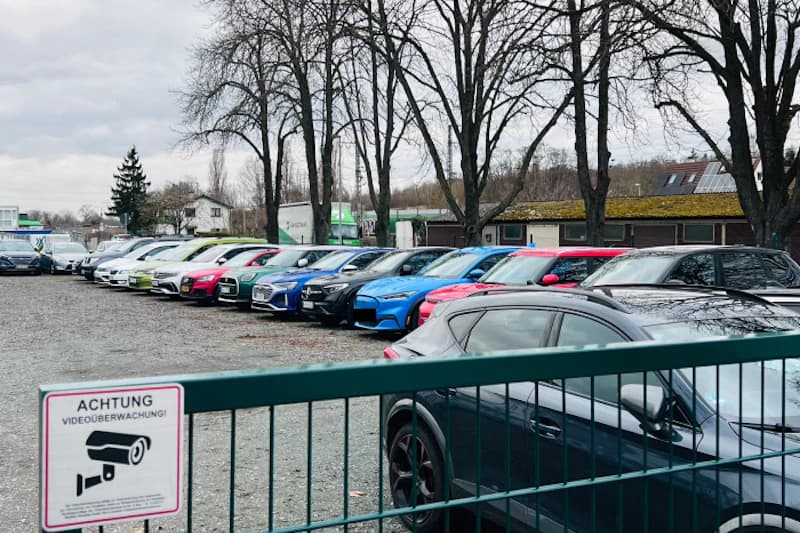 Gitterzaun mit Schild Achtung Videoüberwachung, dahinter Reihe bunt lackierter Autos auf Schotterparkplatz.