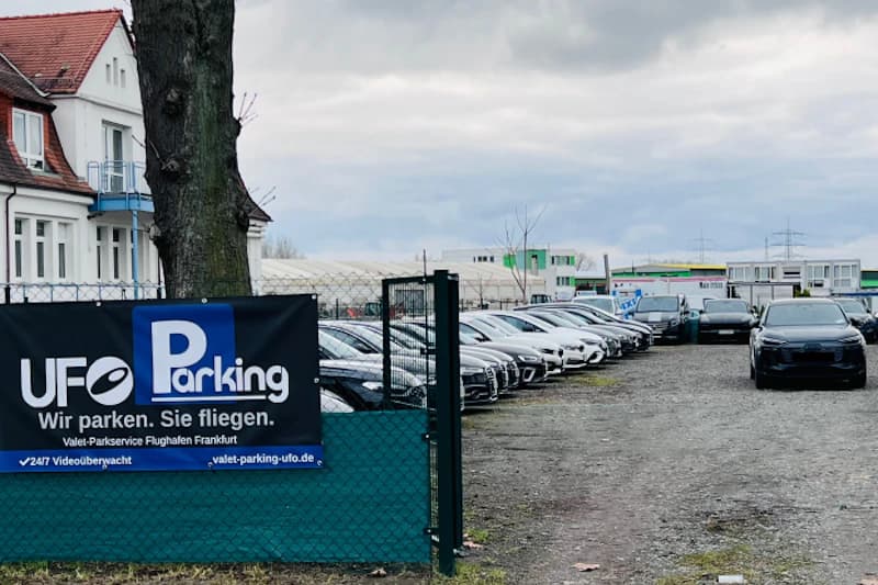Außenparkplatz mit offenem Tor, Banner UFO Parking am Zaun und Reihen geparkter Autos unter bewölktem Himmel.