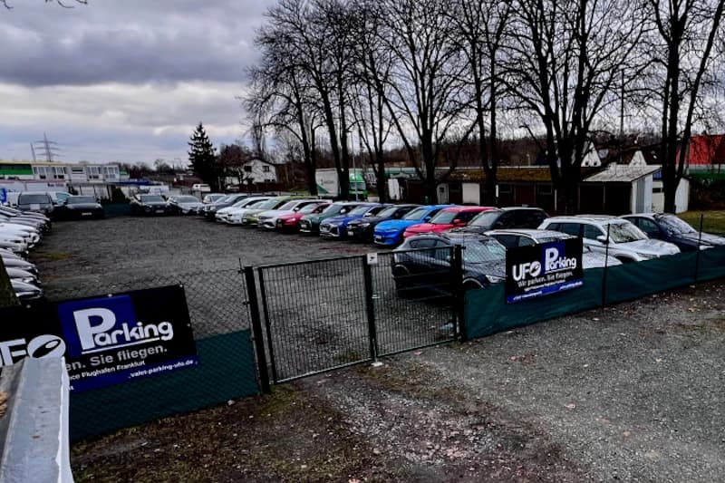Gitterzaun mit geöffneter Einfahrt zu einem Schotterparkplatz mit Reihen geparkter Autos und UFO Parking Werbebanner.