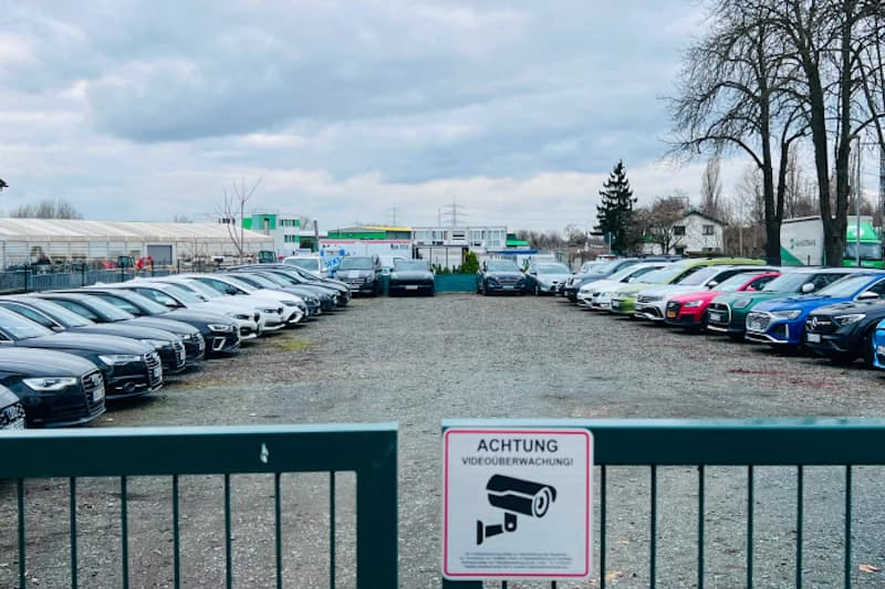 Blick durch Hoftor auf kiesigen Parkplatz mit zwei Reihen geparkter Autos und Schild Achtung Videoüberwachung.