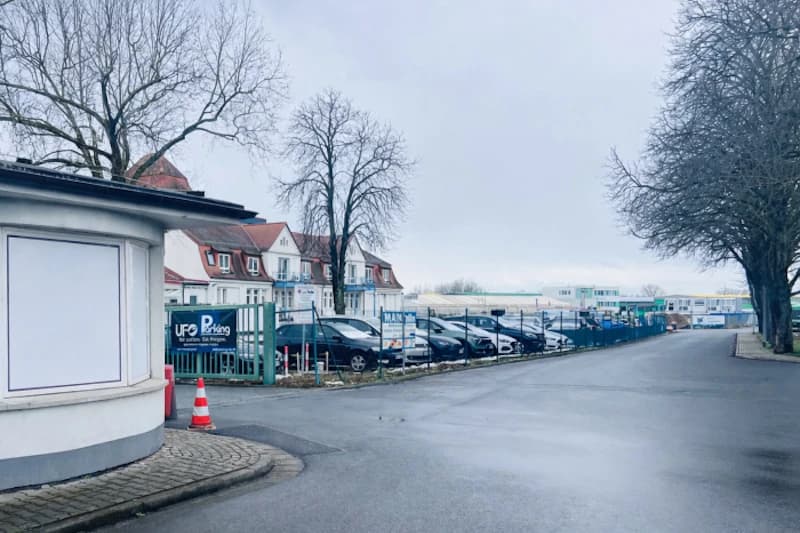 Eingezäunter Parkplatz mit geparkten Autos, UFO Parking Schild, nasse Zufahrtsstraße und winterliche Bäume.