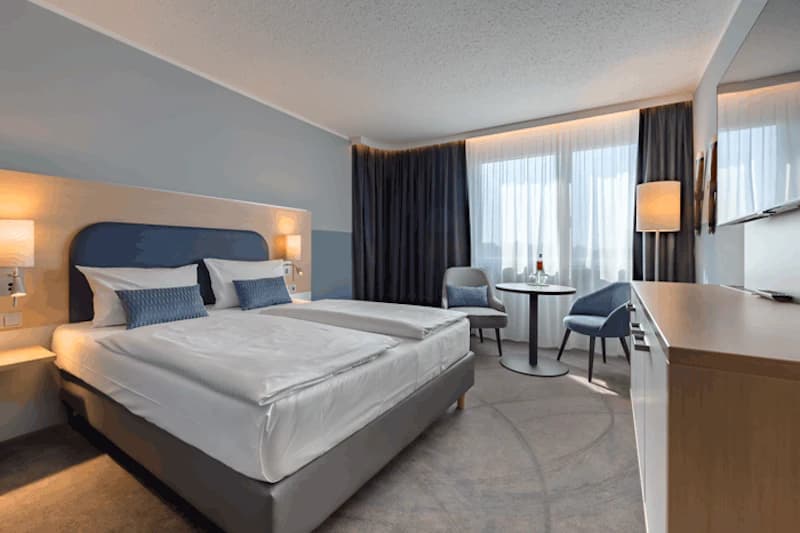 Hotelzimmer mit zwei nebeneinander stehenden Einzelbetten, blauem Kopfteil, Nachttischlampen, Tisch mit zwei Stühlen und Fenster.