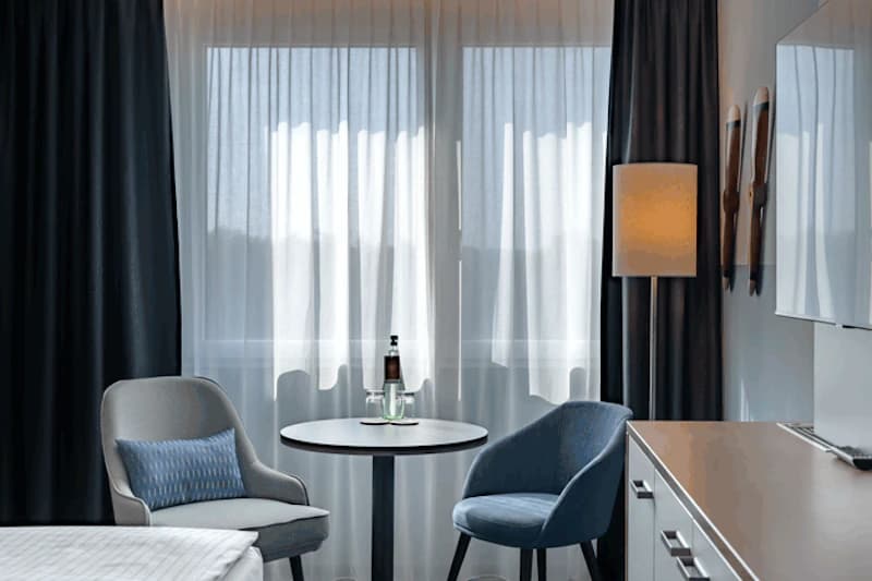 Hotelzimmer mit zwei Sesseln, rundem Tisch mit Flasche und Gläsern, Gardinen, Stehlampe und Sideboard.