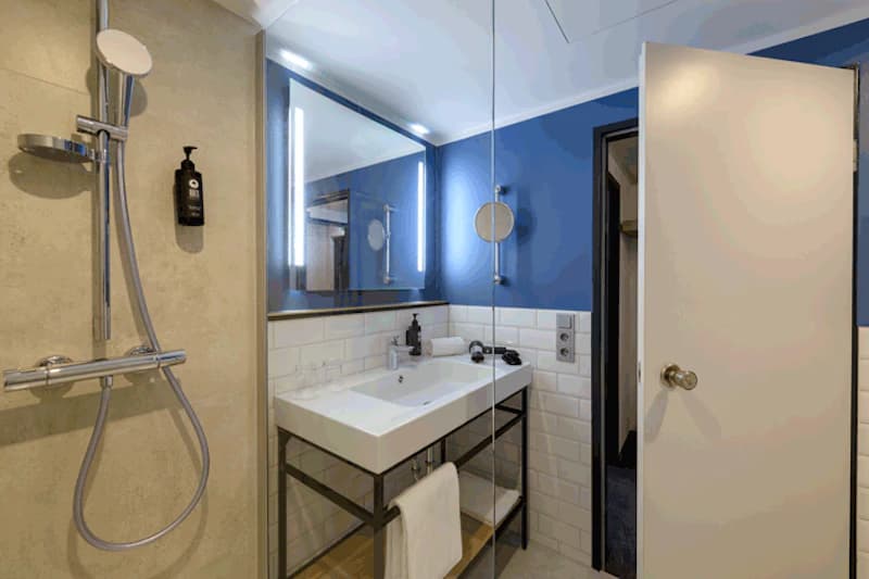 Badezimmer mit Dusche, Handbrause links, Waschbecken, beleuchtetem Spiegel und blau gestrichener Wand.