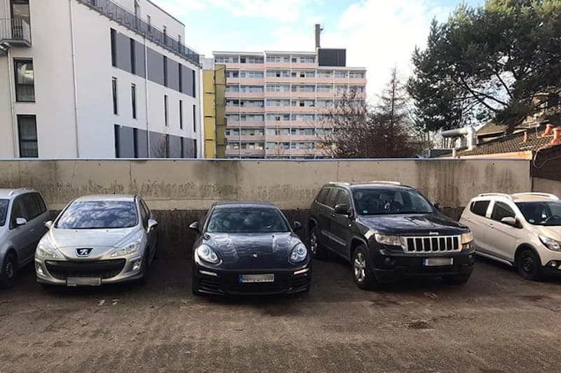 Fünf Fahrzeuge auf Asphaltparkplatz vor niedriger Betonmauer und Wohngebäuden.