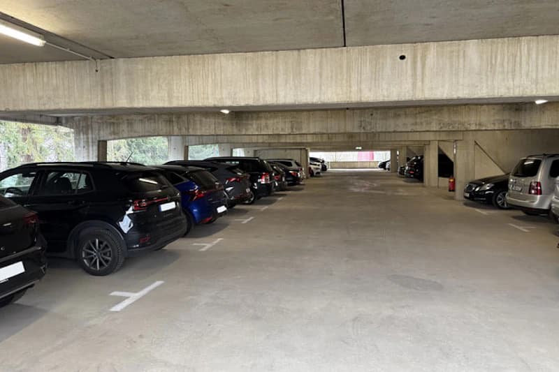 Parkhausinnenraum mit zwei Reihen geparkter Autos, Betonstützen, Deckenbeleuchtung und Feuerlöscher an einer Säule.