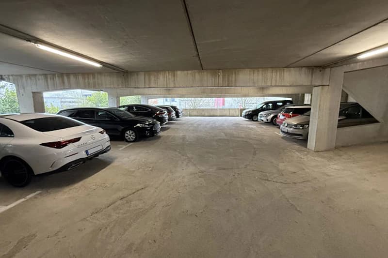 Überdachter Parkhausboden mit geparkten Autos an den Seiten, Betonstützen und Deckenleuchten.