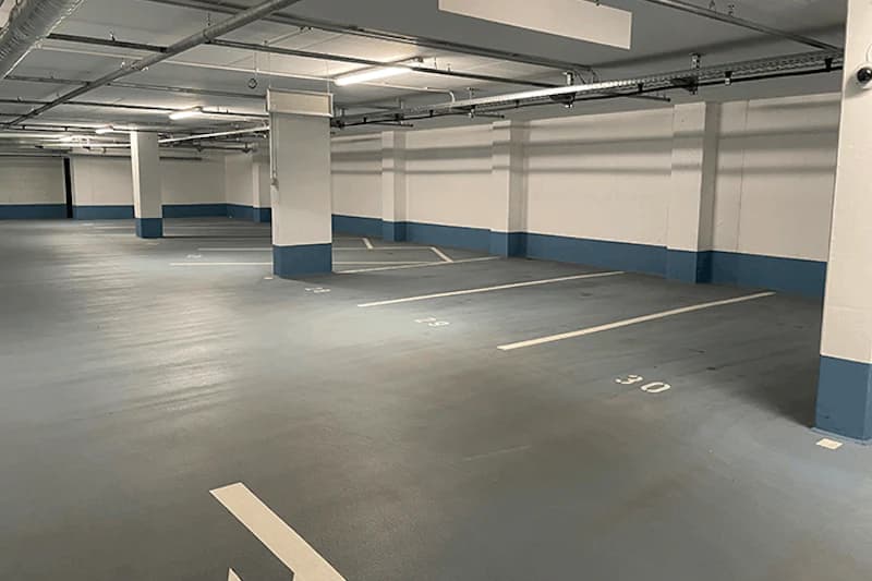 Leeres, gut beleuchtetes Parkhaus mit markierten Stellplätzen, Säulen und Kameras an der Decke.