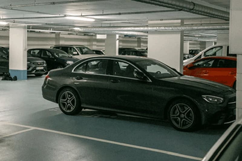 Parkhaus mit Deckenbeleuchtung, Stützpfeilern und mehreren parkenden Autos, im Vordergrund dunkelgraue Limousine.