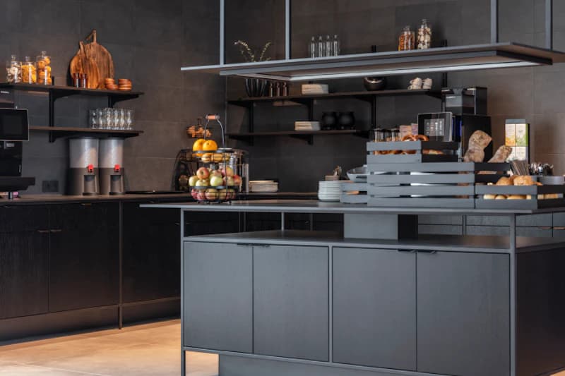 Dunkle Hotelfrühstückstheke mit Obstkorb, Gebäck, Kaffeeautomaten und offenen Regalen.